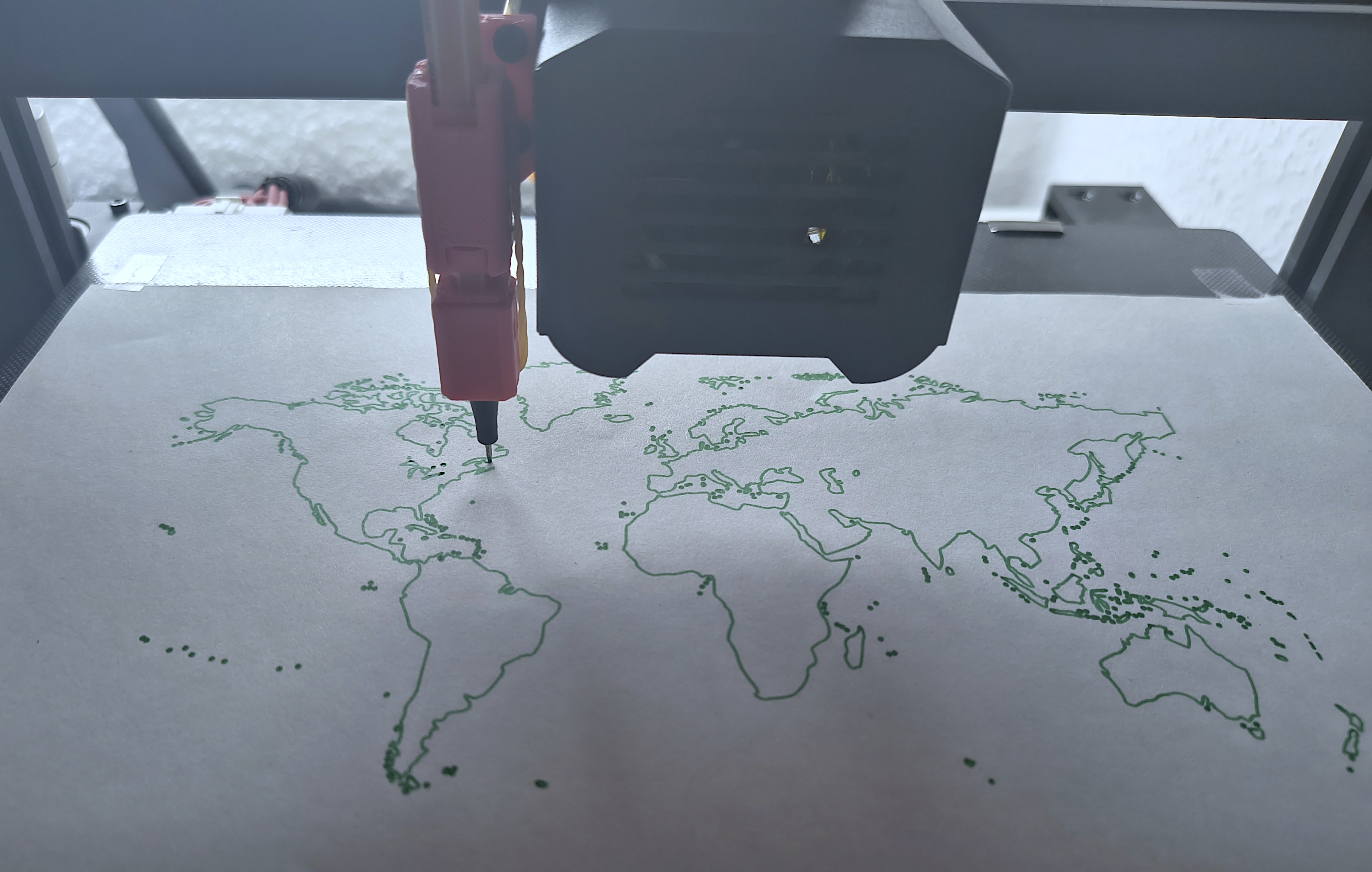 Picture of a 3D printer with a pen-holder mounted. The mounted green pen currently draws a world map.